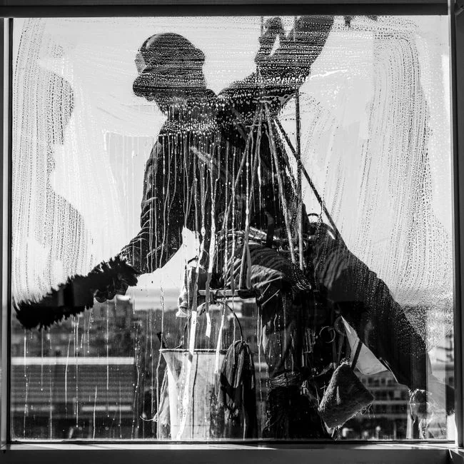 Close-up view of a hand cleaning a window outdoors with a squeegee while wearing yellow gloves.